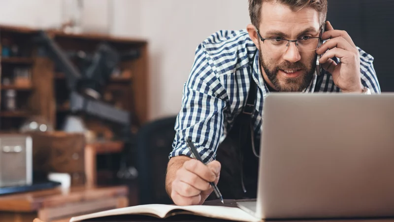 Ein Mann schaut auf einen Laptop mit dem Handy am Ohr und schreibt etwas in ein Notizbuch.