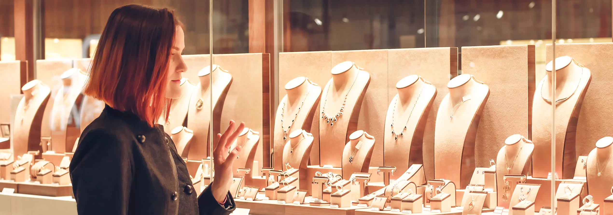 Woman with red hair standing in front of a jewelry store window display, browsing gold necklaces and rings.